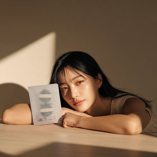 Woman resting her head on a table holding RE LUMI puff package under warm sunlight, showing natural glow and eco-friendly K-beauty charm.